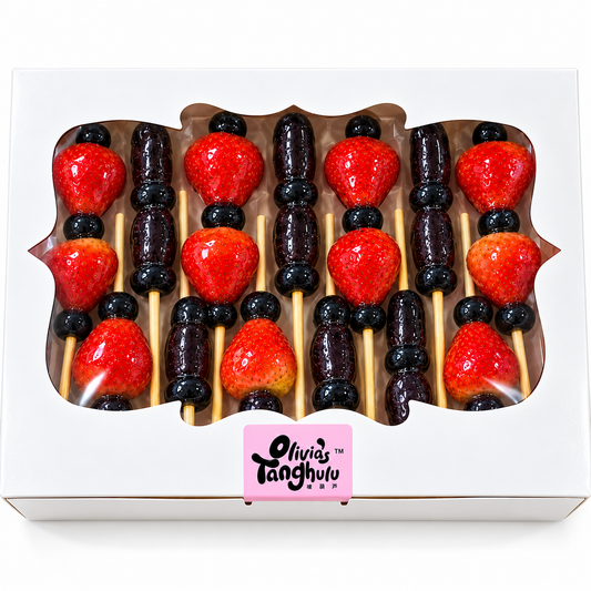 A white gift box containing six skewers of fruit, alternating between bright red strawberries and dark purple-black blueberries, displayed under a clear window on a white background. A pink label at the bottom of the box reads 'Olivia's Tanghulu'.