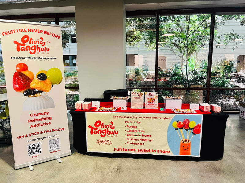 A promotional display booth for Olivia's Tanghulu stands in an indoor event space with large windows showing outdoor greenery. The setup includes a tall white branded banner on the left, a black-clothed display table with packaged fruit skewers, and a large front sign with product examples and event usage text.